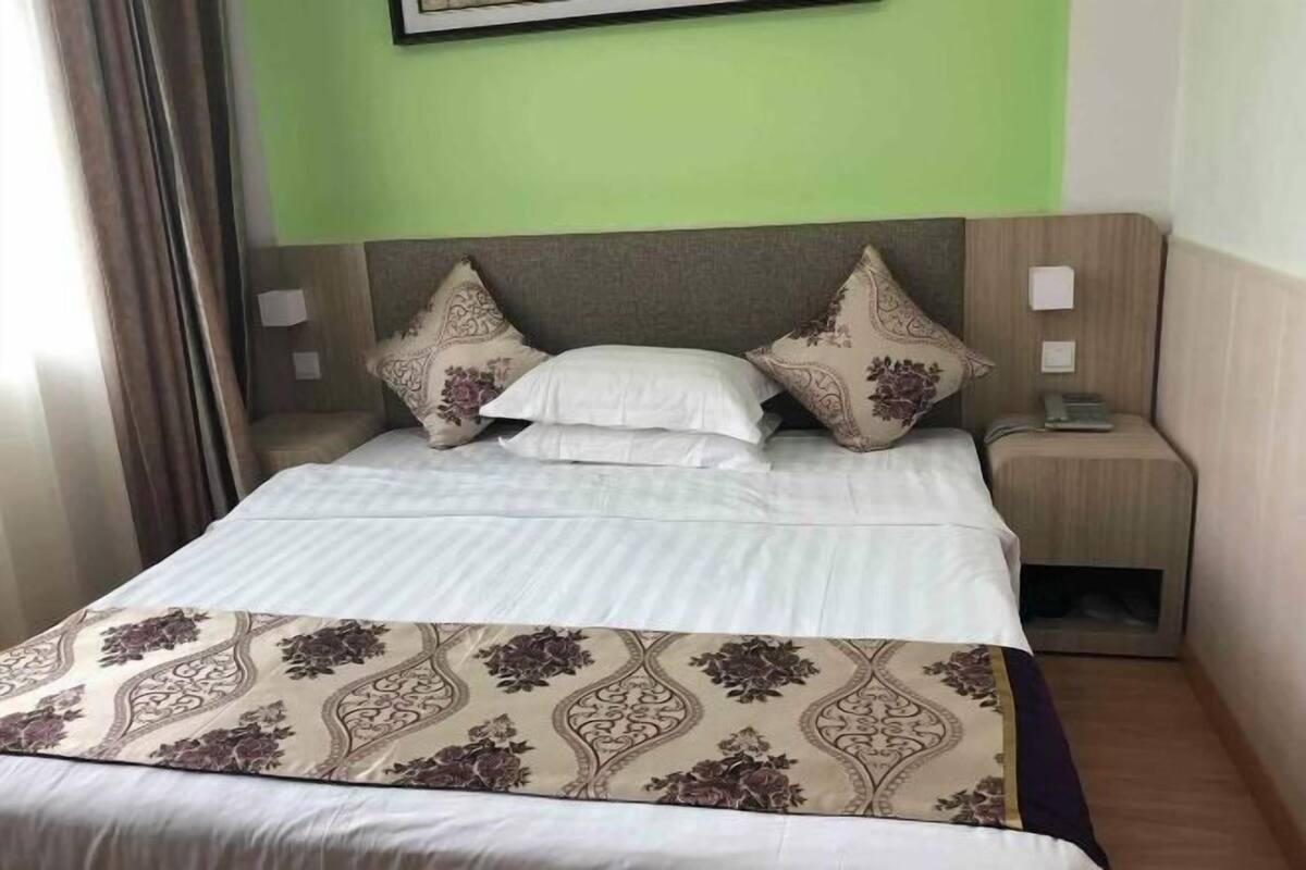 A neatly made bed with a patterned bedspread, a headboard with decorative pillows, a bedside table with a lamp, and a wall-mounted phone.