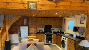 Un acogedor interior de cabaña de madera con una sala de estar que incluye un sofá y una mesa de comedor, una cocina con armarios de madera y electrodomésticos modernos, y una escalera que conduce a un nivel superior.