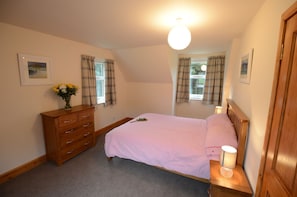 A bedroom with a bed, a dresser, a nightstand with a lamp, and two windows with curtains.