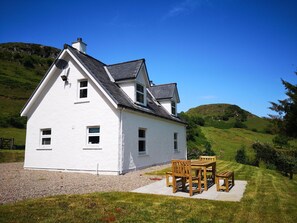 A white house with a sloped roof, a chimney, and a small porch with wooden furniture. - Achalic Beag - sleeps 4 guests  in 2 bedrooms (Oban)