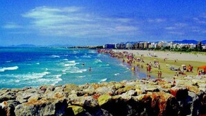 A beach with rocky foreground, clear blue water, and a crowded beach area with buildings in the background. - Relax at Mallorca Villa Cas Padri Jordi (Muro)