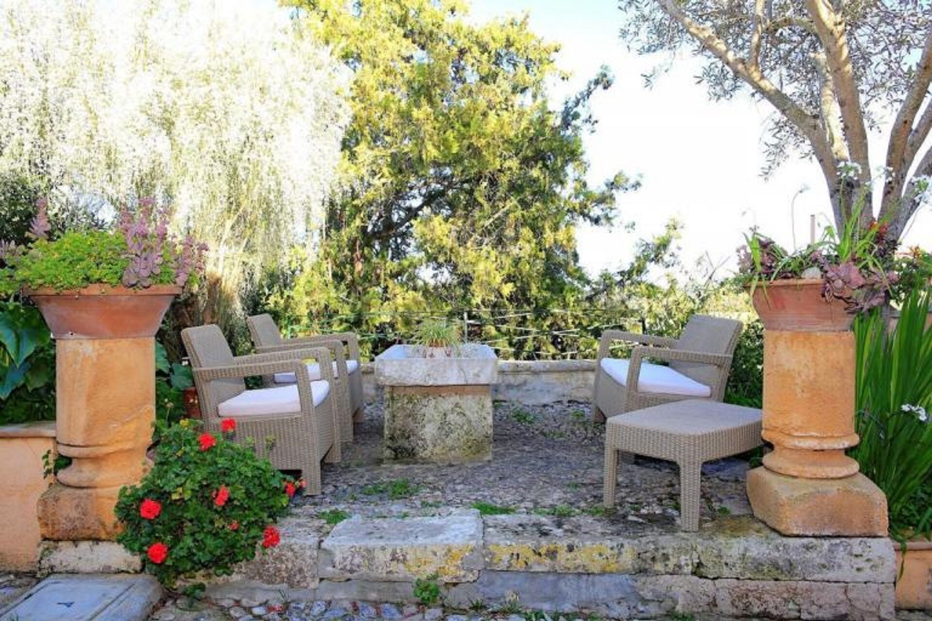 An outdoor seating area with wicker chairs and a stone table, surrounded by potted plants and a stone wall.