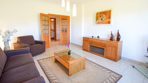 Ein Wohnzimmer mit einem braunen Sofa, einem Glastisch und einem Holzschrank. Zwei Türen führen in andere Räume.