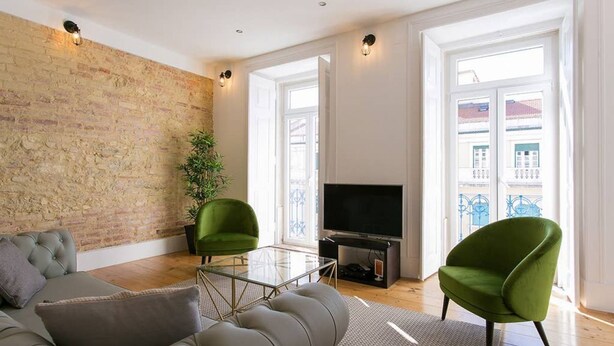 Una sala de estar moderna con pared de ladrillo, un sofá, dos sillones verdes, una mesa de centro de vidrio y un televisor de pantalla plana.