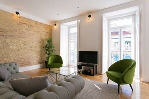 A modern living room with a brick wall, a sofa, two green armchairs, a glass coffee table, and a flat-screen TV.