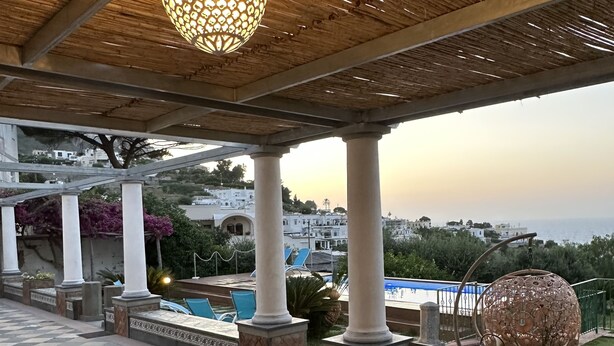 A covered outdoor area with a tiled floor, a hanging light fixture, and a view of a pool and buildings in the background. - Villa Amalu Capri private pool (Capri)