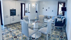 A dining area with a glass table surrounded by white chairs, a white sofa with blue and white cushions, and a kitchen with white cabinets and a built-in oven. - Villa Amalu Capri private pool (Capri)