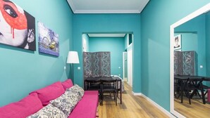Une pièce aux murs de couleur turquoise, avec un canapé rose, un plaid à motifs, une table à manger et des chaises, ainsi qu’un paravent décoratif.
