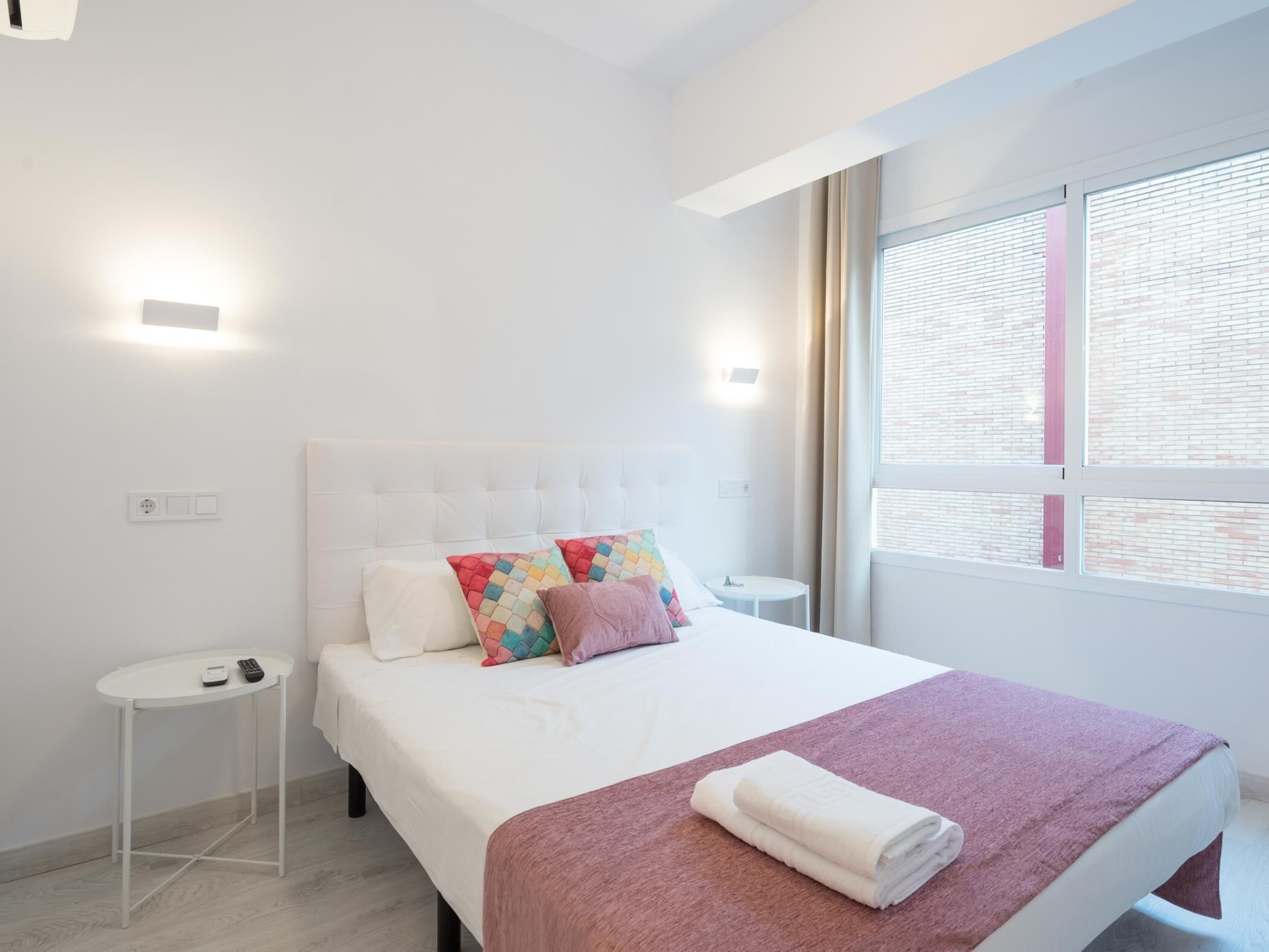 Ein modernes Hotelzimmer mit einem ordentlich bezogenen Bett, einem kleinen Nachttisch mit Telefon und einem Fenster mit Jalousien.