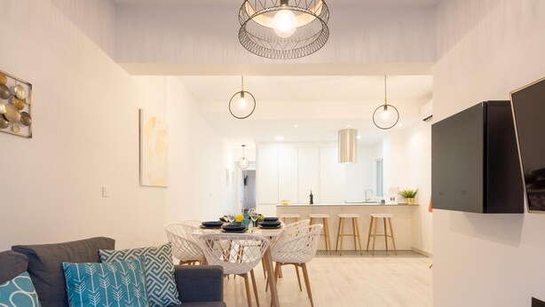 A modern living room with a grey sofa, patterned cushions, a dining area with a white table and chairs, and a kitchen in the background. - New Center 3Bedrooms 2 Bath Apartment (Malaga)