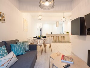 Un salon moderne avec un canapé gris, des coussins à motifs, un coin repas avec une table et des chaises blanches, et une cuisine en arrière-plan.
