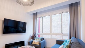 A modern living room with a flat-screen TV mounted on the wall, a sofa, a small round table with a book and remote, and a potted plant. - New Center 3Bedrooms 2 Bath Apartment (Malaga)