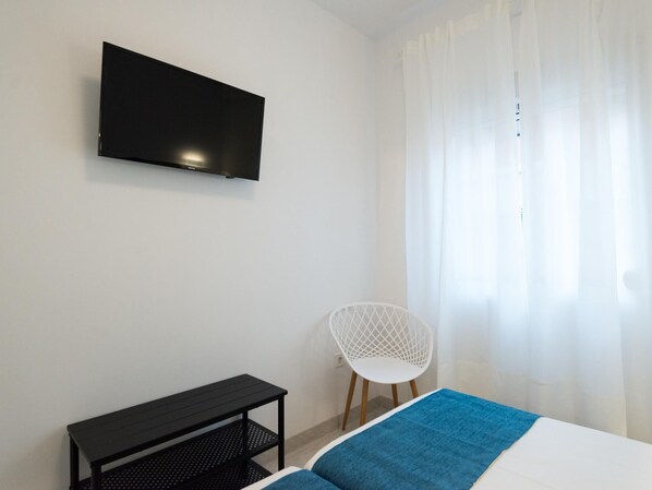 Une chambre à coucher avec un lit, un téléviseur fixé au mur, une petite table noire, une chaise blanche et une fenêtre avec des rideaux légers.