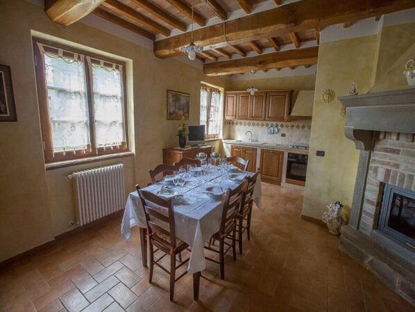 Un coin repas avec une table dressée pour six personnes, des chaises en bois, une cheminée et une cuisine en arrière-plan.