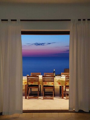 An open doorway leading to a dining area with wooden chairs and a table, overlooking a body of water during sunset.
