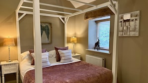 Een hemelbed met een bruine sprei, twee nachtkastjes met lampen, een raam met jaloezieën, en een schilderij aan de muur.