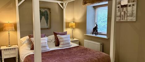 A four-poster bed with a brown quilt, two bedside tables with lamps, a window with blinds, and a painting on the wall.