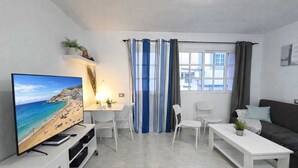 Ein modernes Wohnzimmer mit Flachbildfernseher, weißen Möbeln, einem Fenster mit Vorhängen und Blick auf einen Strand.