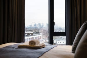 A bed with a rolled towel and a view of a cityscape through a window with curtains. - Deluxe Victoria Home close to Buckingham Palace (London)