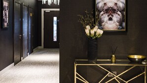 En hall med mörka väggar, en modern lampa, ett inramat konstverk, en vas med blommor och ett konsolbord med guldfärgad ram, där det står en skål och ett ljus.