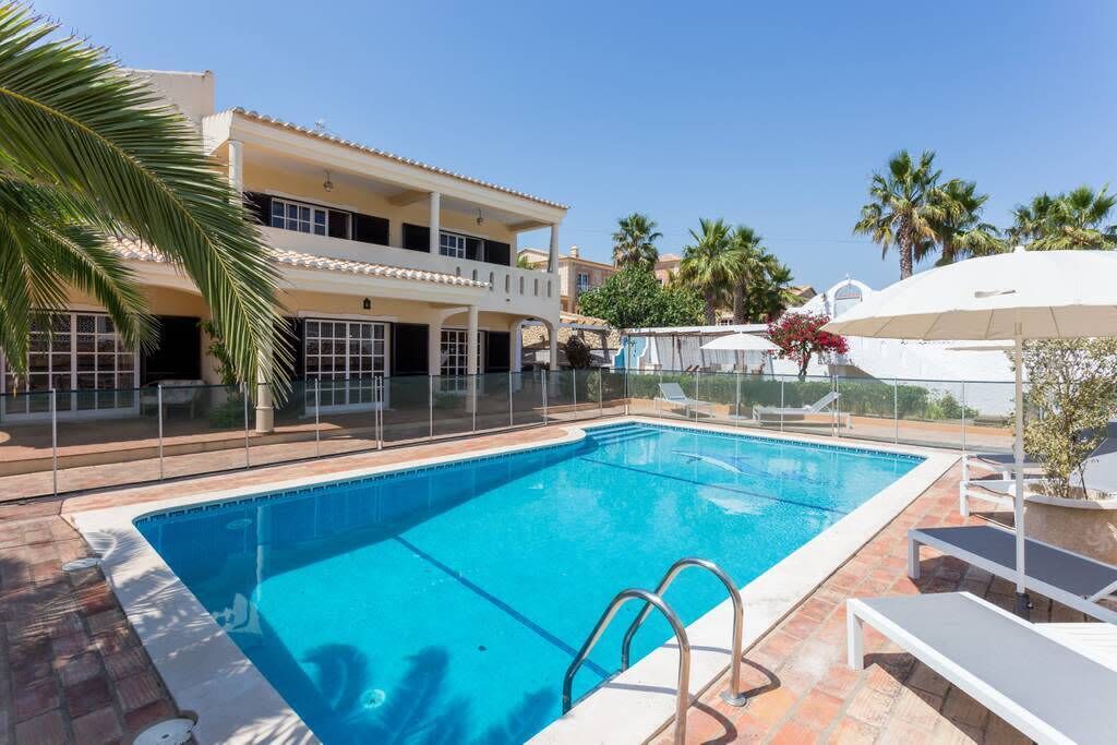 A swimming pool with a clear blue water, surrounded by a white fence, and a building with balconies and windows in the background.