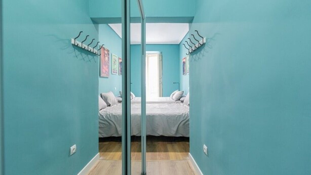 Een smalle gang die leidt naar een slaapkamer met teal muren, een houten vloer en een bed met witte beddengoed.