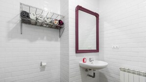Een badkamer met witte betegelde wanden, een witte wastafel met rode omlijsting, een metalen handdoekrek met handdoeken, een toilet en een afvalbak.