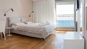 Un dormitorio moderno con cama, mesitas de noche, un escritorio y una silla. Tiene un balcón con barandilla y vistas al mar.