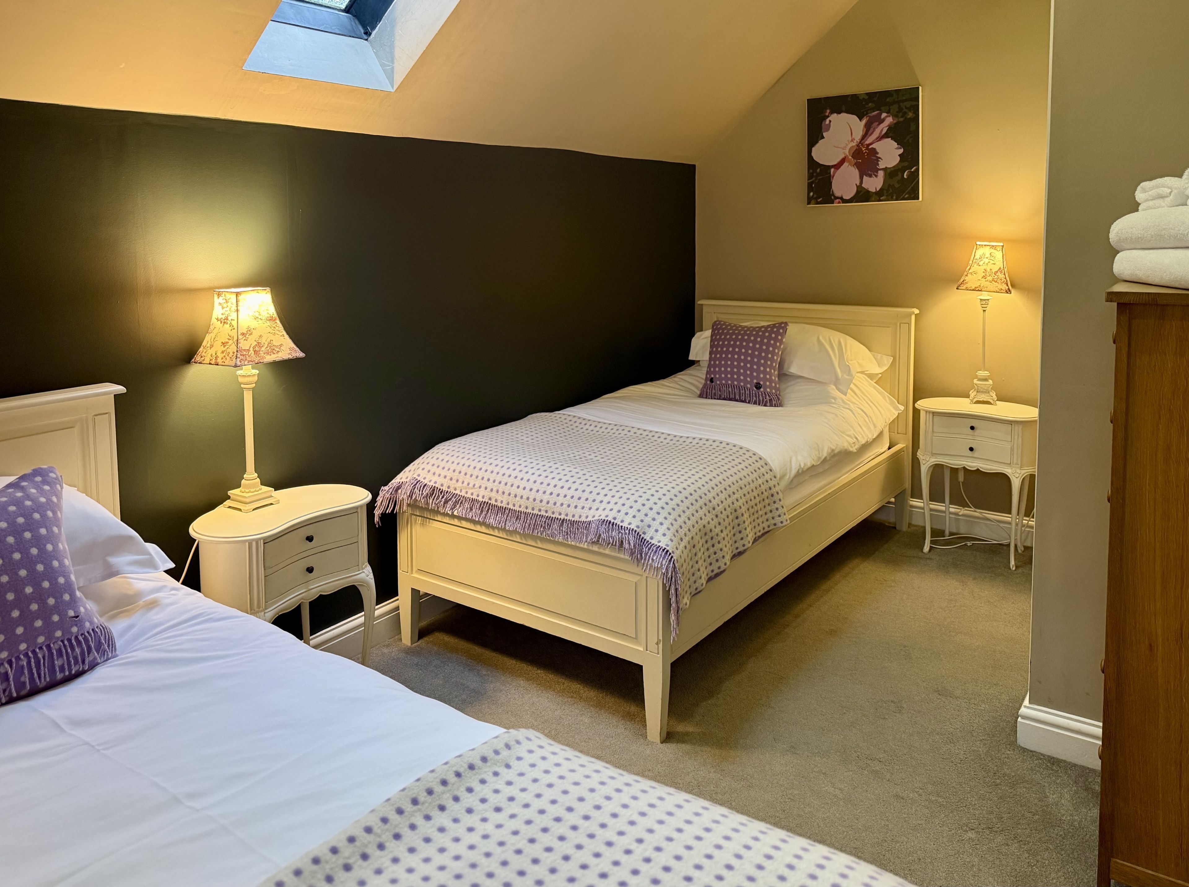 A bedroom with two beds, a nightstand with a lamp, a bedside table with a lamp, and a painting on the wall.