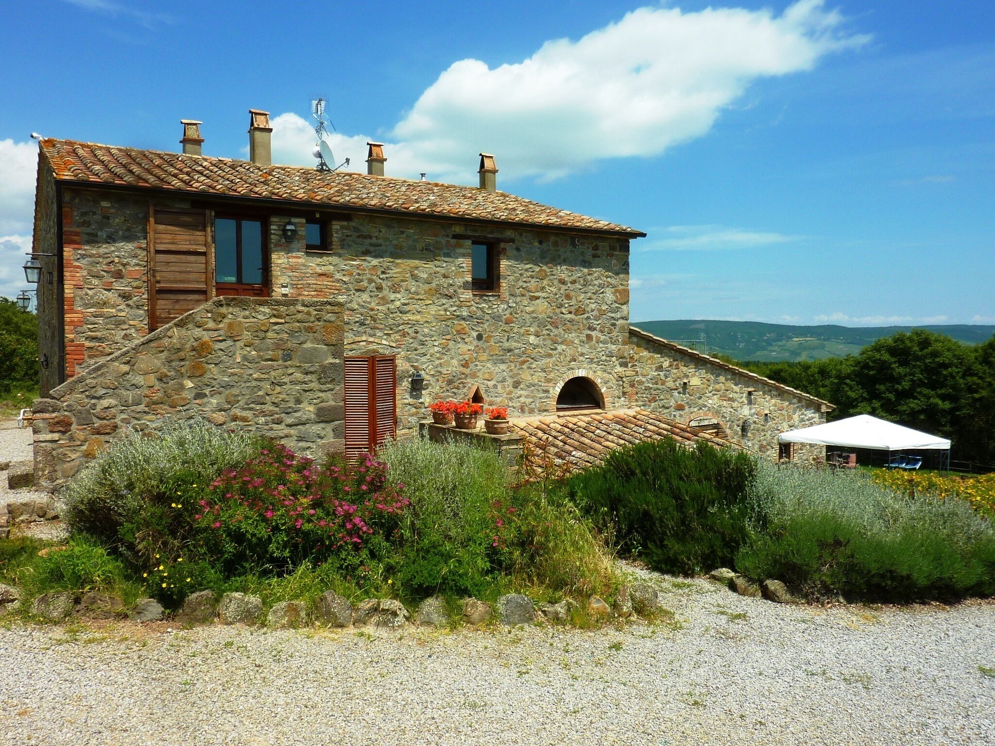 Una casa de piedra con techo de tejas, un pequeño jardín con flores y vistas al campo.