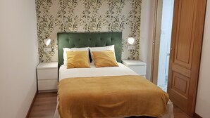 Ett sovrum med en grön, klädd sänggavel, två sänglampor, en säng med ett senapsgult överkast och ett blommigt tapetmönster.