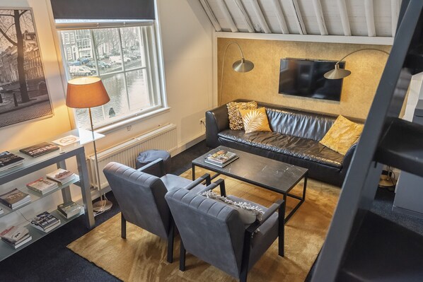 A modern living room with a sofa, coffee table, and chairs. There is a bookshelf with books and a lamp. A window offers a view of a canal with boats. - Spaarne Luxury Residence Haarlem (Haarlem)