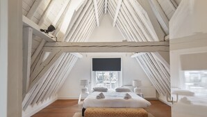 Un dormitorio con techo inclinado, un ventanal grande, una cama con almohadas, un banco y dos mesitas de noche con lámparas.