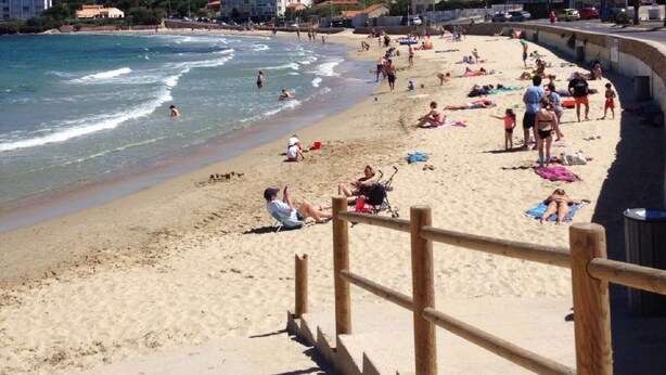 Ein Strand mit sonnenbadenden Personen und Schwimmern, eine Holzumzäunung und Gebäude im Hintergrund.