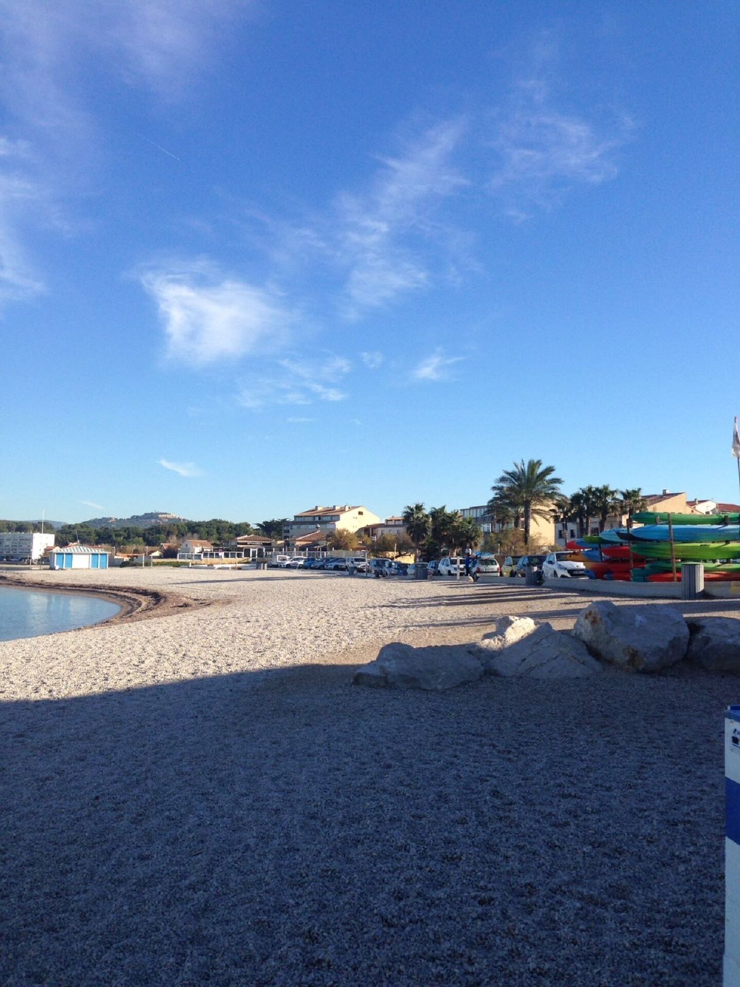A beach with pebbles, a small building, and a row of parked cars.