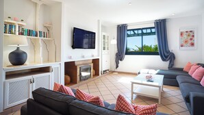 A living room with a fireplace, sofa, TV, and bookshelves. - Casa Nicole, Macher, Free heated Pool, High Speed Wifi and Scenic views Sleeps 8 (Macher)