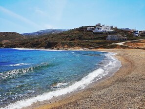 A coastal landscape with clear blue waters, a pebbly beach, and white buildings on a hillside.