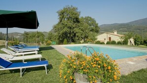 Un espace piscine doté de chaises longues et d’un parasol, entouré de verdure et bordé d’un bâtiment en pierre.