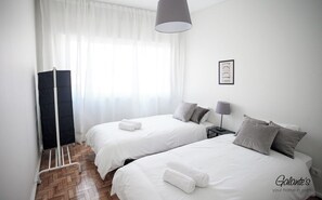 A hotel room with two beds, a bedside table with a lamp, a window with sheer curtains, and a framed picture on the wall.