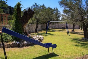 Children can play on the playground equipment, including a seesaw, swing set, and climbing structure, surrounded by trees and grass.