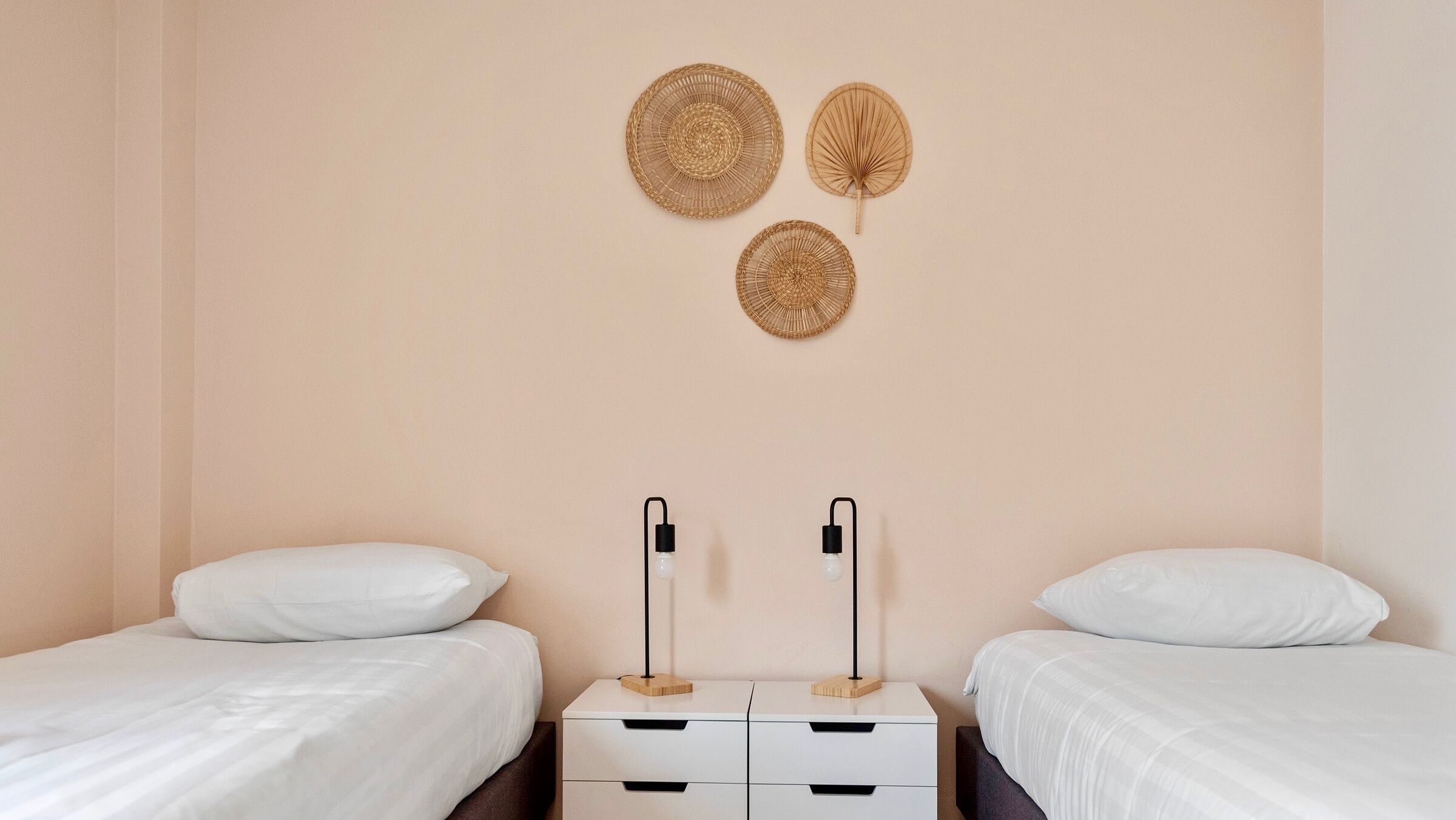 A room with two single beds, a small white dresser with drawers, and three woven wall decorations.