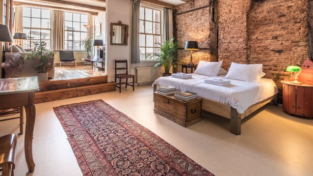 Ein rustikales Schlafzimmer mit unverputzten Backsteinwänden, einem Holzbett, einem großen Fenster und Blick auf die Stadt.