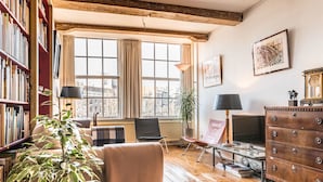 Ein gemütliches Wohnzimmer mit Holzboden, Kamin, Bücherregal, bequemen Sessel und einem großen Fenster mit Blick auf die Stadt.