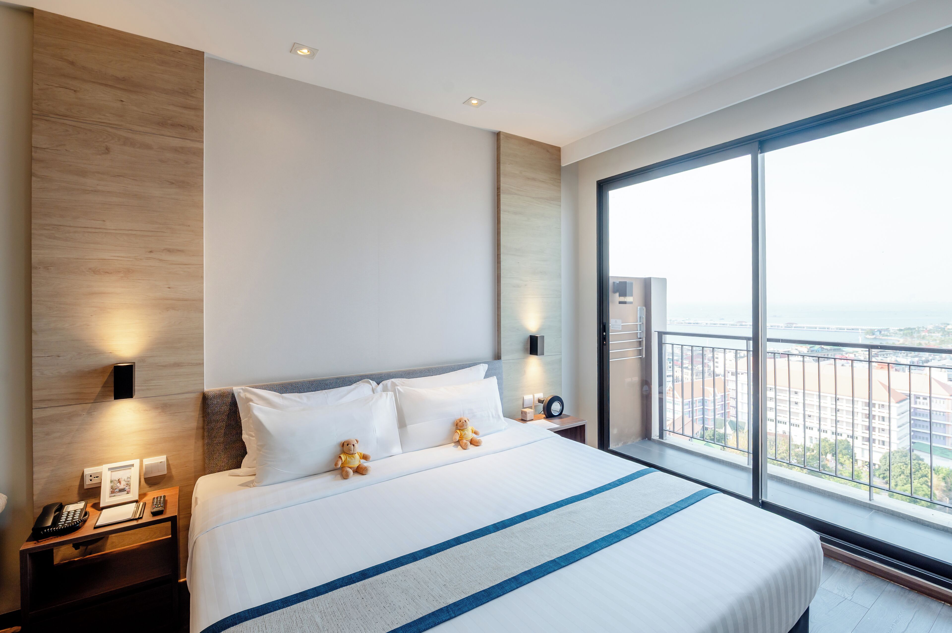 A hotel room with a large bed, two bedside lamps, a nightstand with a phone and a picture frame, and a view of the city through large windows.