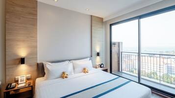 A hotel room with a large bed, two bedside lamps, a nightstand with a phone and a picture frame, and a view of the city through large windows.