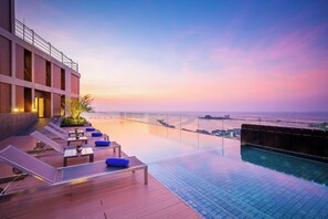 Una piscina infinita en la azotea con sillones de descanso y vistas del horizonte urbano al atardecer.