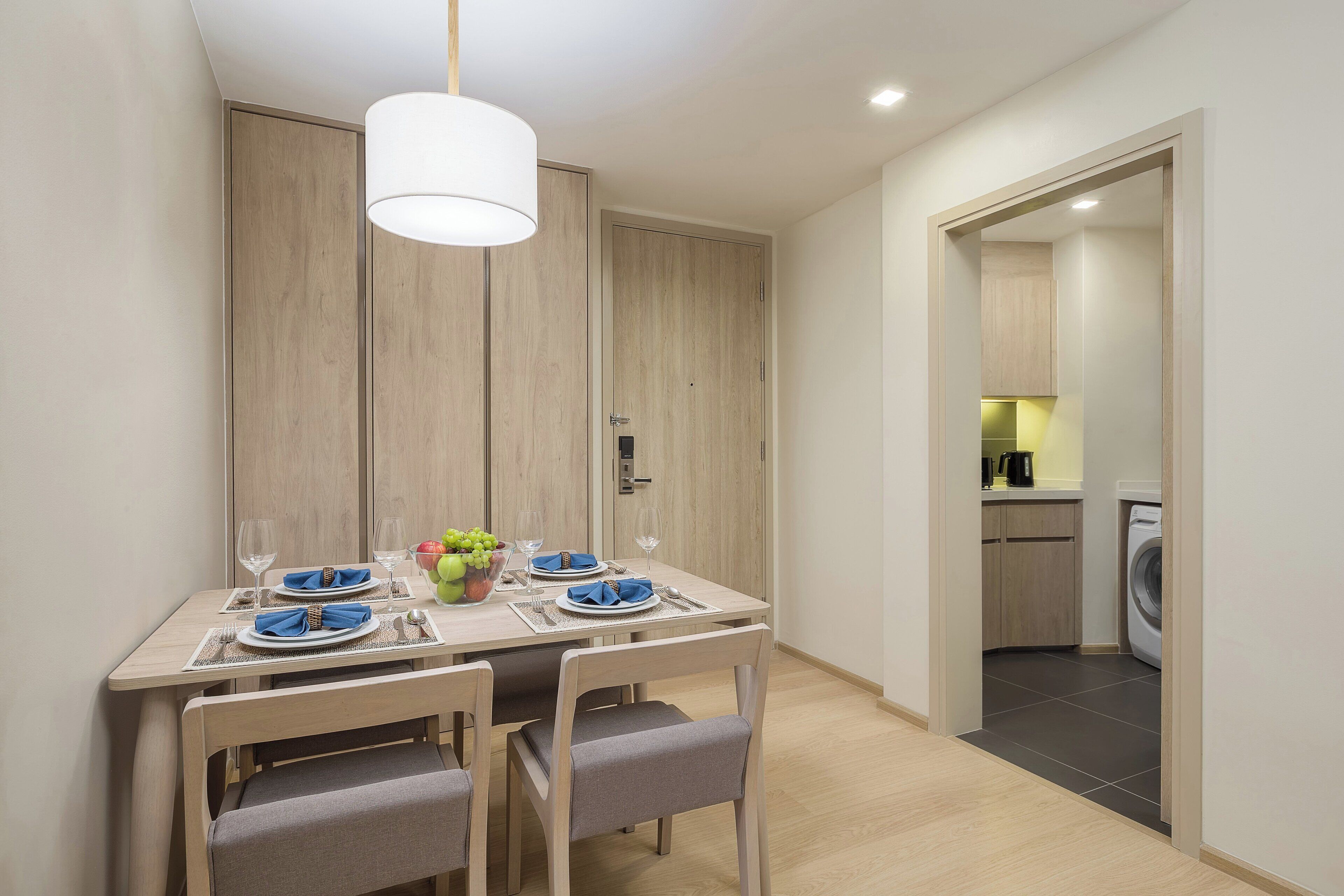 A dining area with a table set for four, a washing machine in the background, and a kitchen visible through an open door.