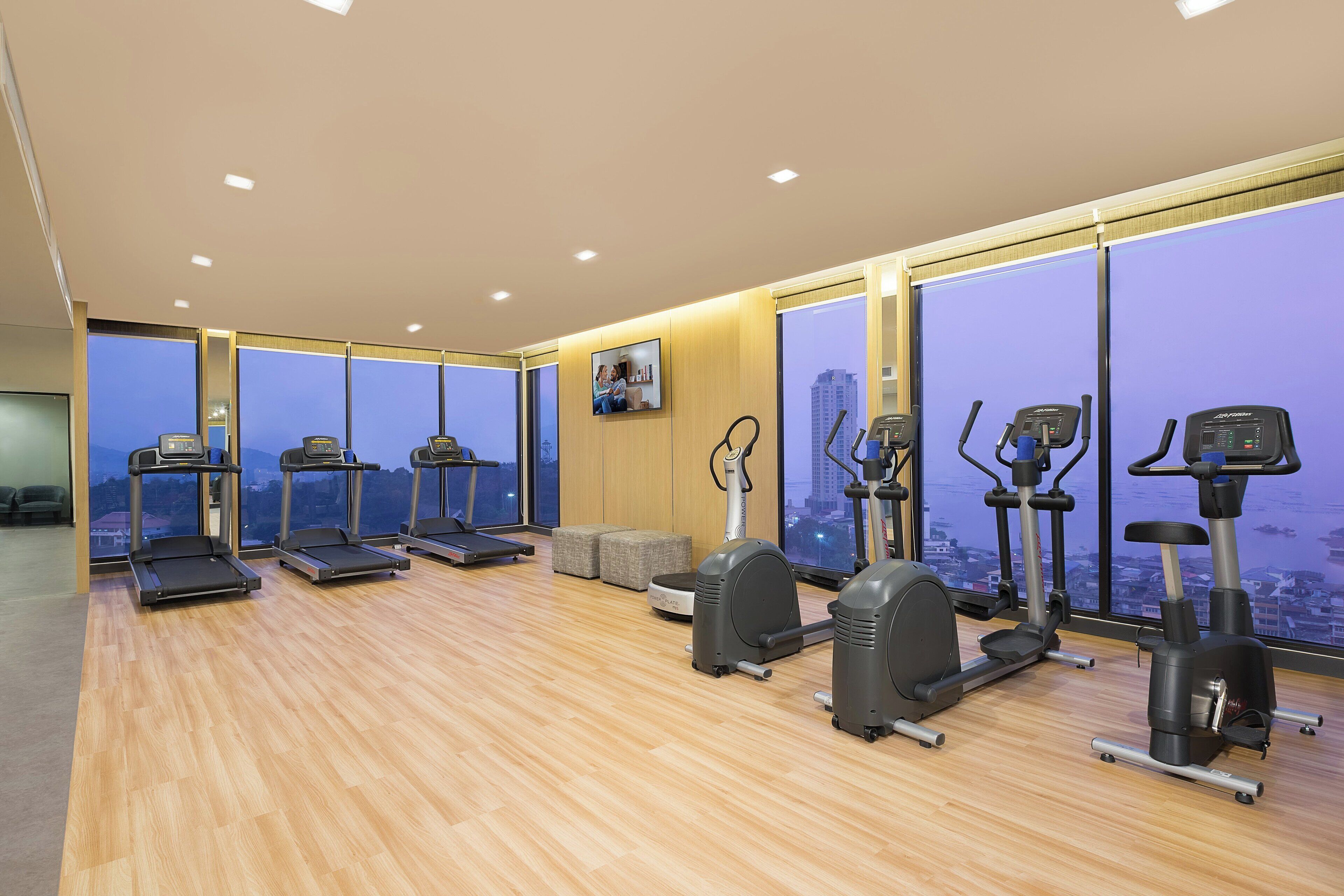A modern gym with treadmills, elliptical machines, and a rowing machine. It features large windows with a city view.
