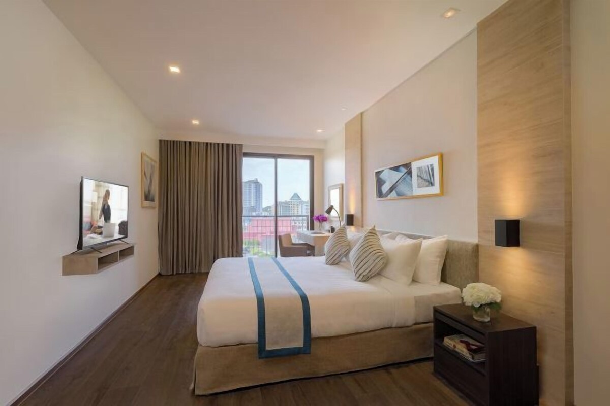 A modern hotel room with a large bed, a desk, a chair, a TV, and a view of the city through the window.