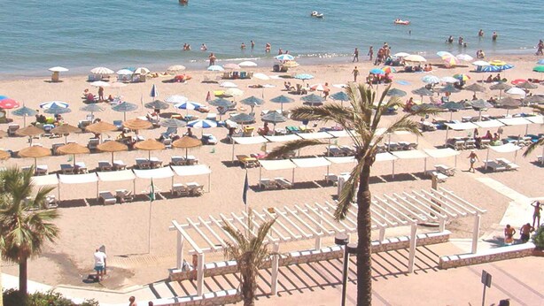 Een strand met veel parasols en ligbedden, een heldere blauwe zee en enkele mensen in het water.